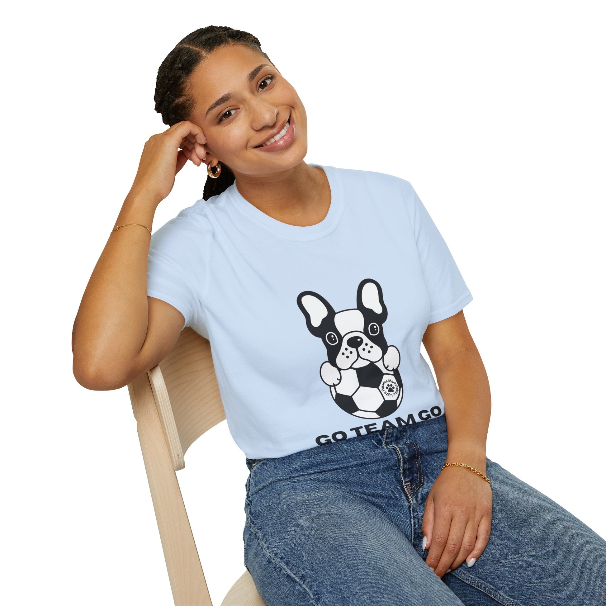 A woman in a white shirt with a dog print sits on a chair, showcasing the Soccer Dog Unisex T-Shirt. The tee, made of soft 100% cotton, features twill tape shoulders and a ribbed collar for durability and comfort.