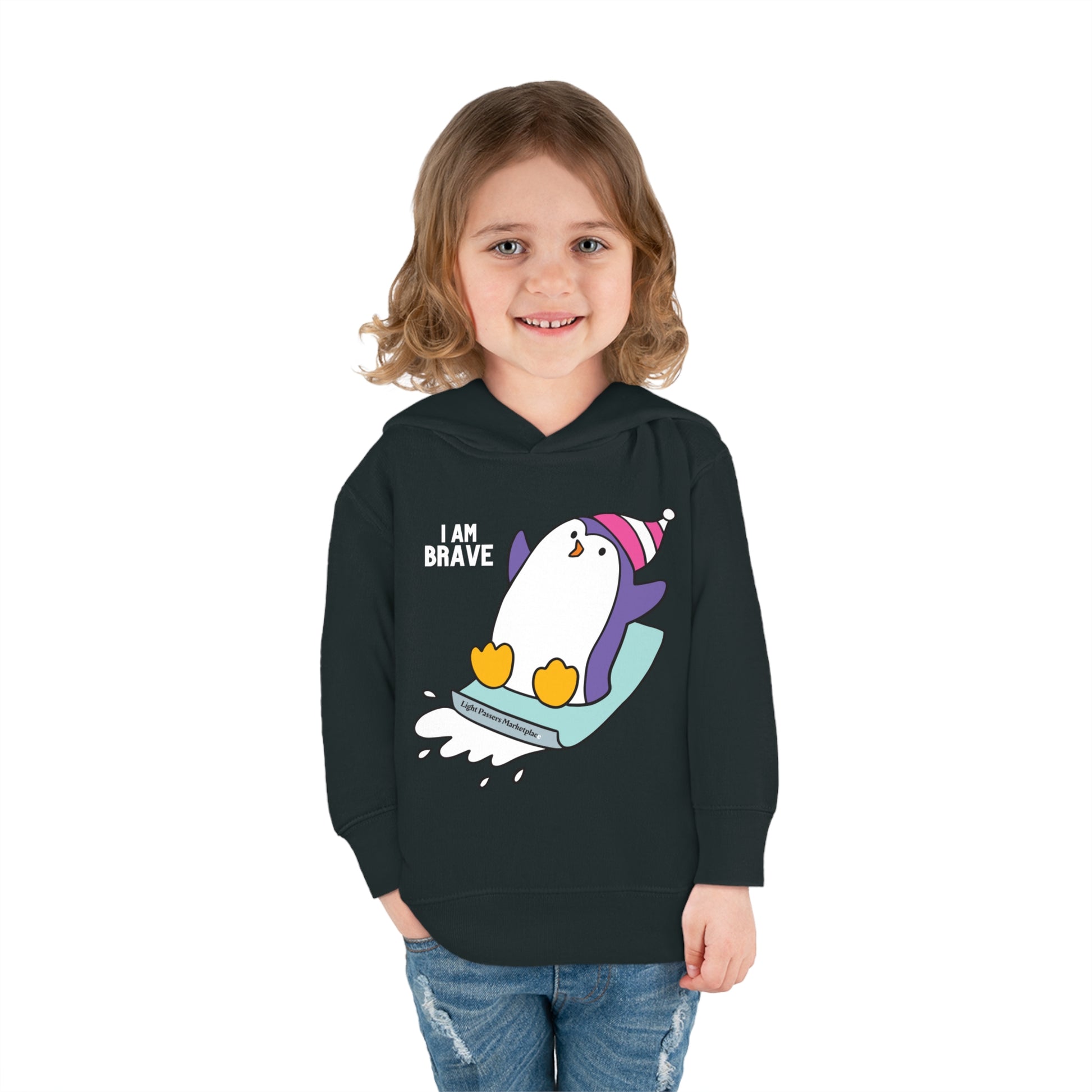 A child in a Brave Penguin Toddler Hooded Sweatshirt, smiling. Black sweatshirt with penguin design, cartoon penguin on snowboard visible. Jersey-lined hood, side seam pockets for cozy wear.