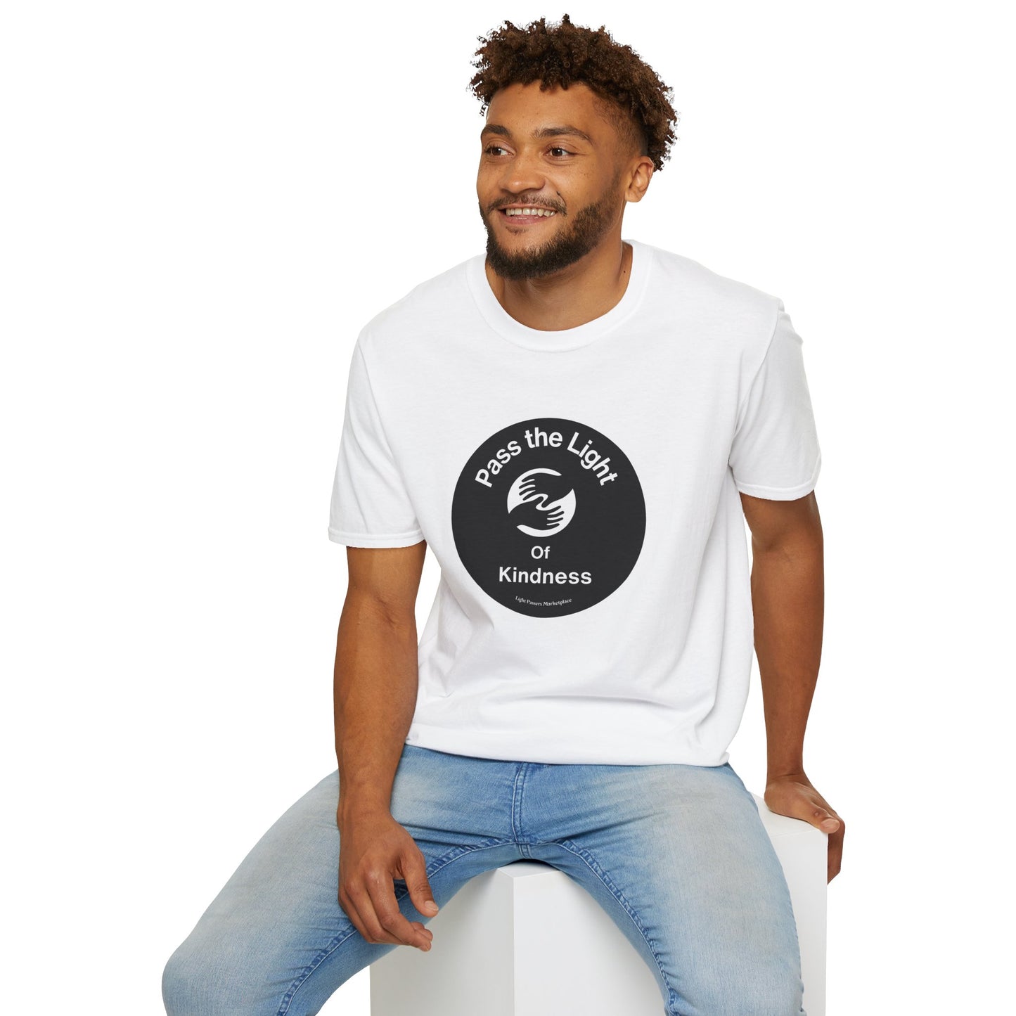 A man in a white shirt sits on a cube, showcasing the Pass the Light of Kindness 2 hands Unisex T-shirt. The heavy cotton tee offers a classic fit with smooth fibers for vivid printing.