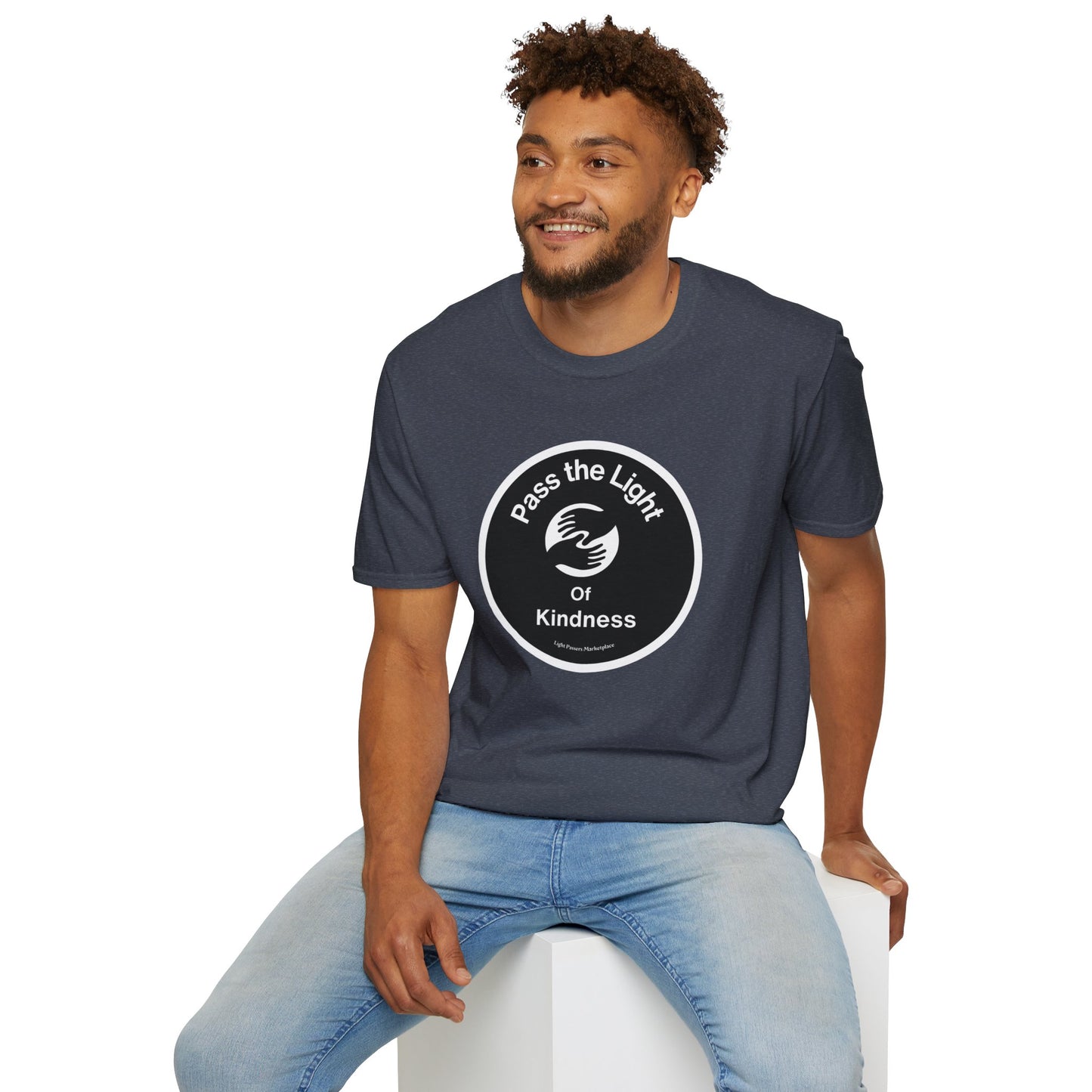 A man in a Pass the Light of Kindness 2 hands Unisex T-shirt, sitting on a cube, with a black and white sticker nearby. Tee features smooth fabric, no side seams, and shoulder tape for durability.