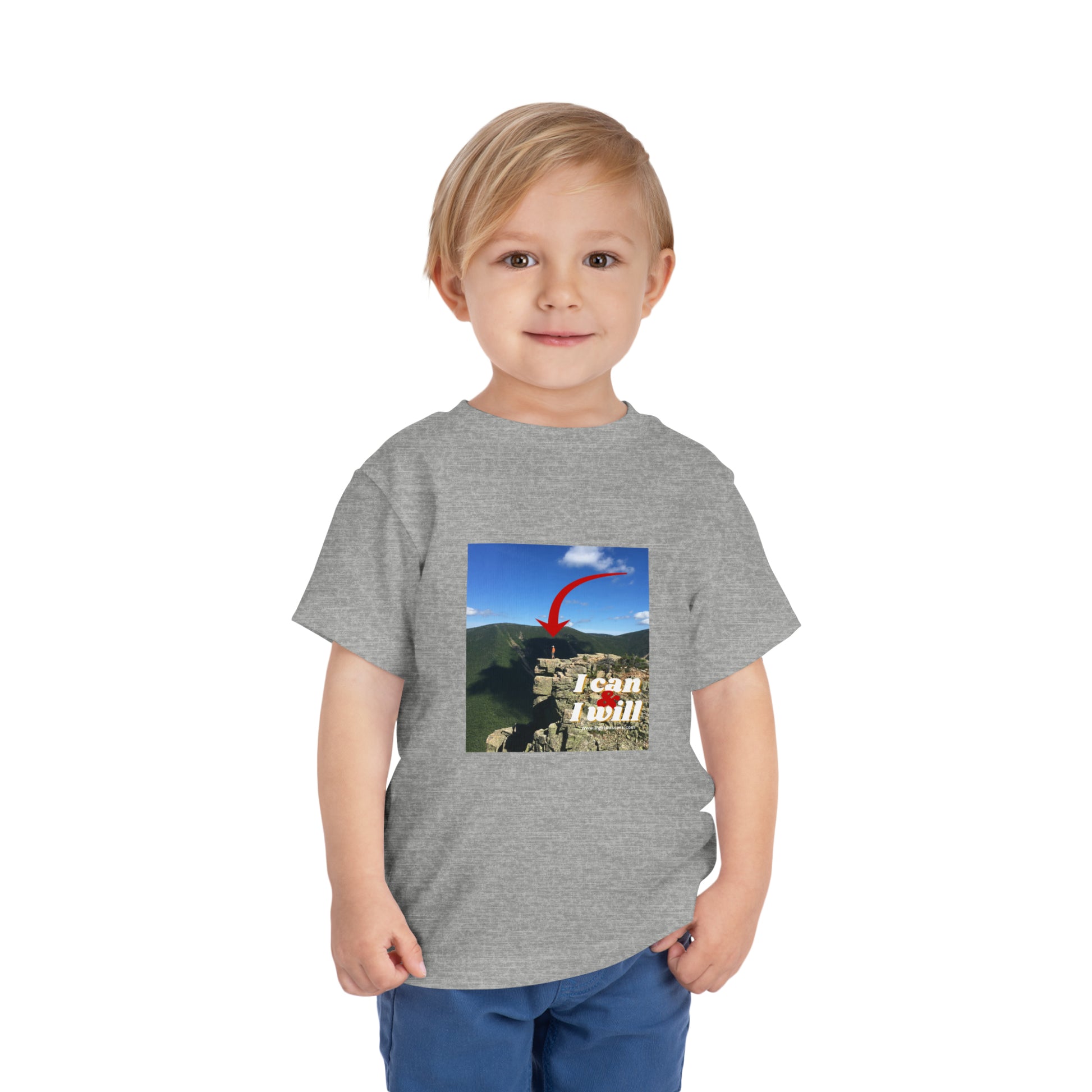 A toddler wearing a grey I Can and I Will hiking t-shirt, featuring a child's face close-up and a person standing on a cliff. Made of 100% Airlume combed cotton.