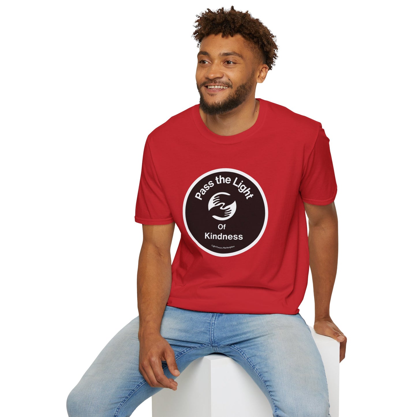 A man in a red shirt sits on a cube, showcasing Pass the Light of Kindness 2 hands Unisex T-shirt. The image features a person in casual attire, emphasizing the classic fit and 100% cotton fabric for premium comfort.