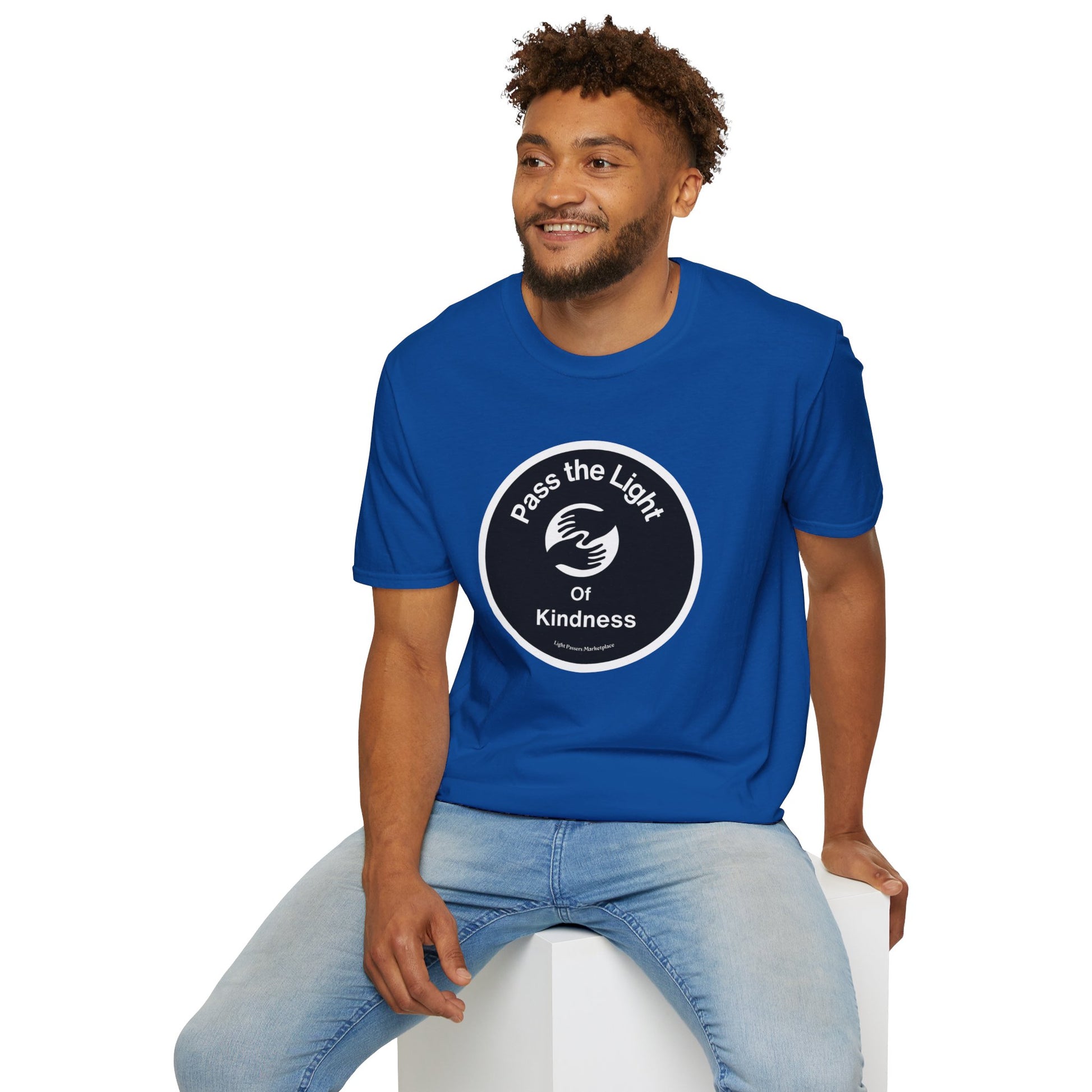 A man in a blue shirt with a white circle and hand design sits on a cube, showcasing the Pass the Light of Kindness 2 hands Unisex T-shirt. Classic fit, 100% cotton tee with no side seams for comfort.