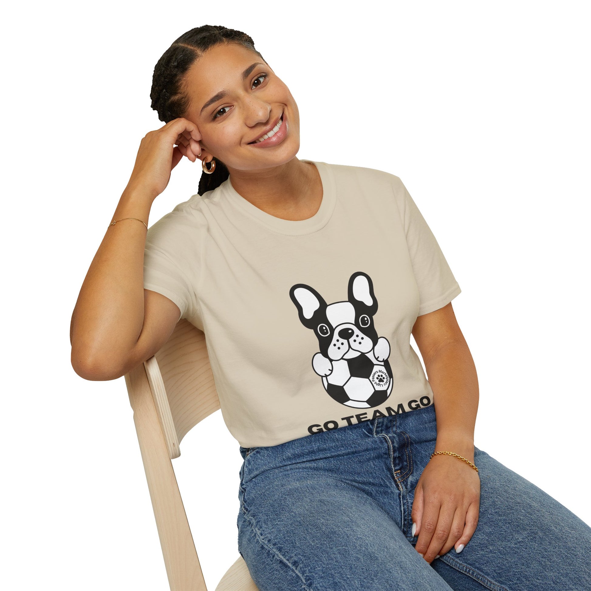 Person wearing Soccer Dog Unisex T-Shirt featuring a cartoon bulldog with a soccer ball and GO TEAM GO slogan, smiling while seated on a wooden chair.