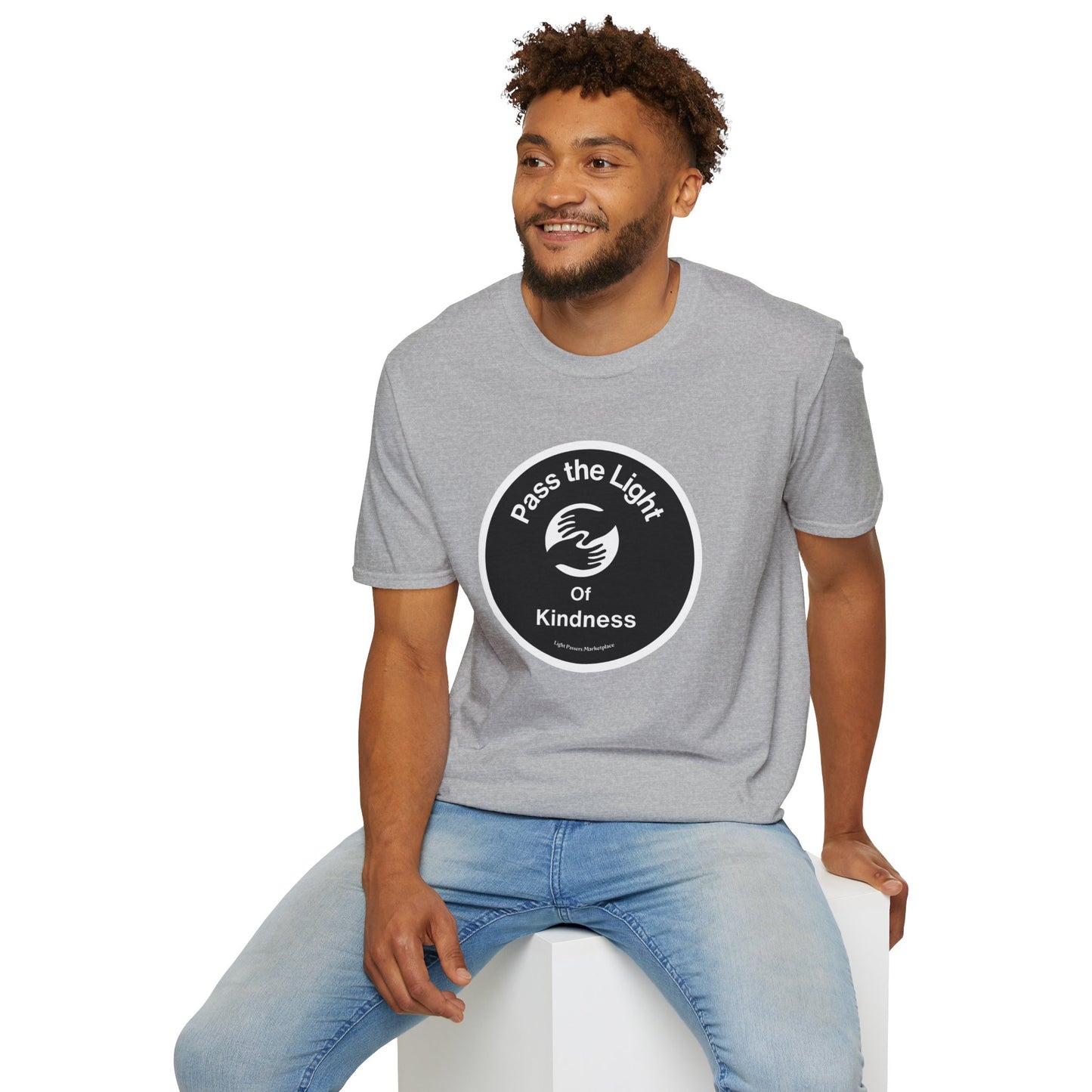 A man in a grey shirt sits on a cube, showcasing the Pass the Light of Kindness 2 hands Unisex T-shirt. Close-up of logo and face details. Cotton tee with smooth surface for vivid printing.