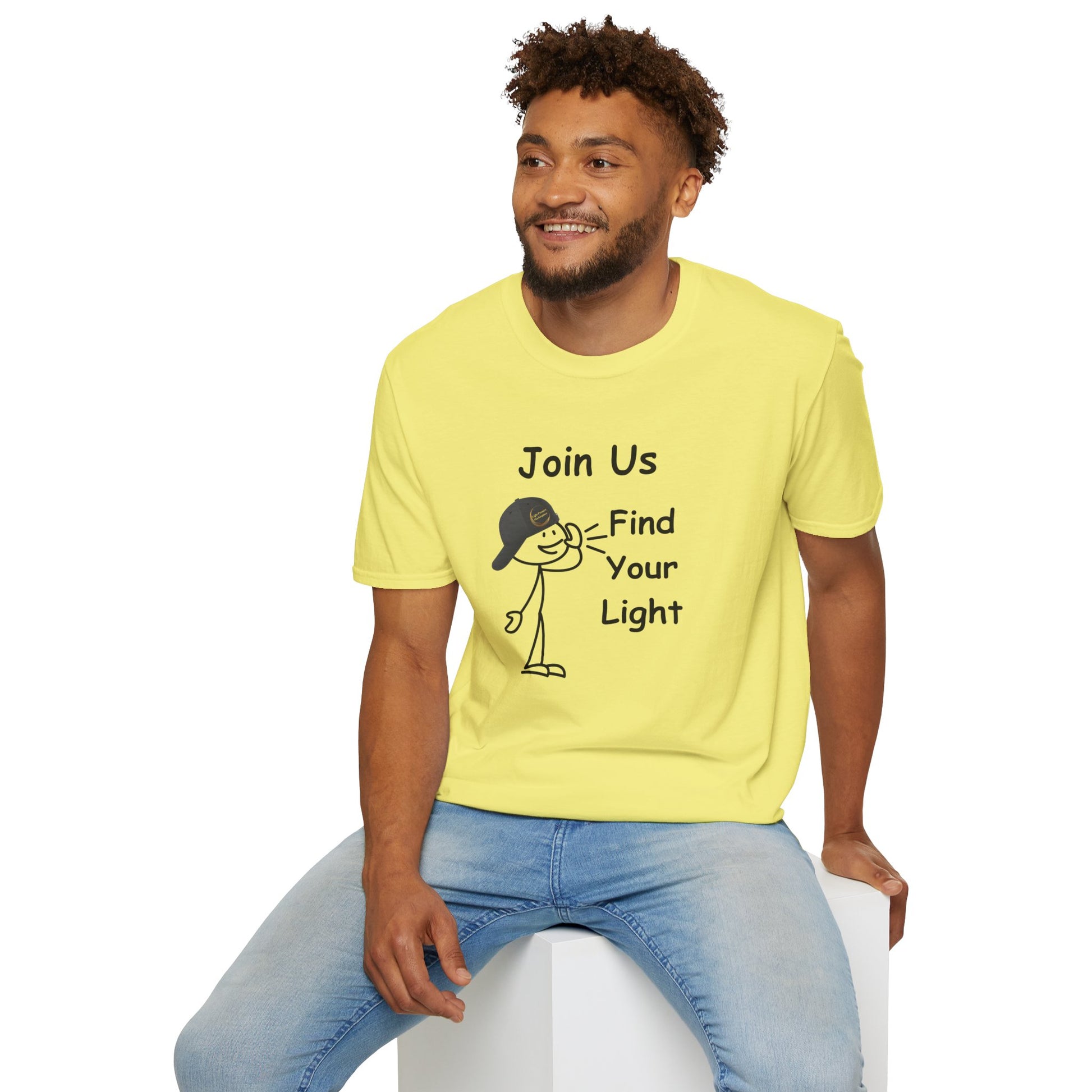 A man in a yellow shirt with a cartoon character and text sits on a cube, showcasing the Join Us Find Your Light Unisex T-Shirt's casual comfort and 100% cotton fabric with twill tape shoulders.