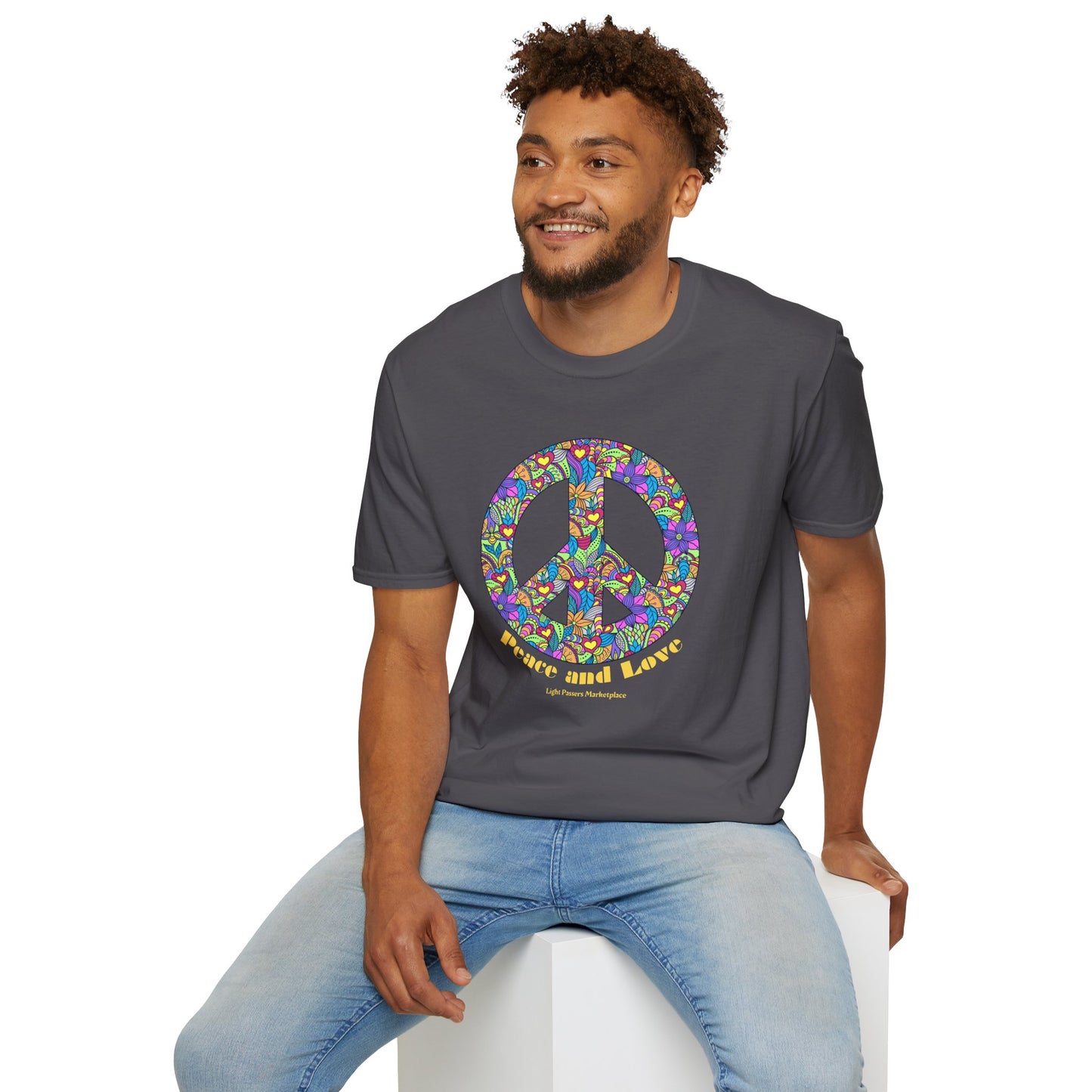 A man in a grey shirt with a colorful peace sign sits on a cube, showcasing the Peace Sign with Flowers Unisex T-shirt. Made of soft 100% cotton, featuring a crew neckline and tear-away label for comfort.