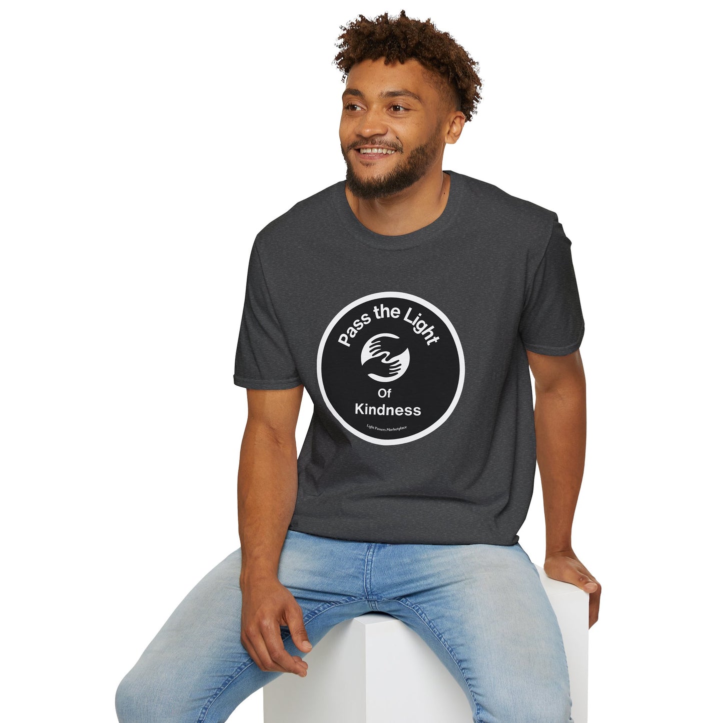 A man in a grey shirt sits on a cube, showcasing the Pass the Light of Kindness 2 hands Unisex T-shirt. The image captures a casual, versatile tee perfect for everyday wear.