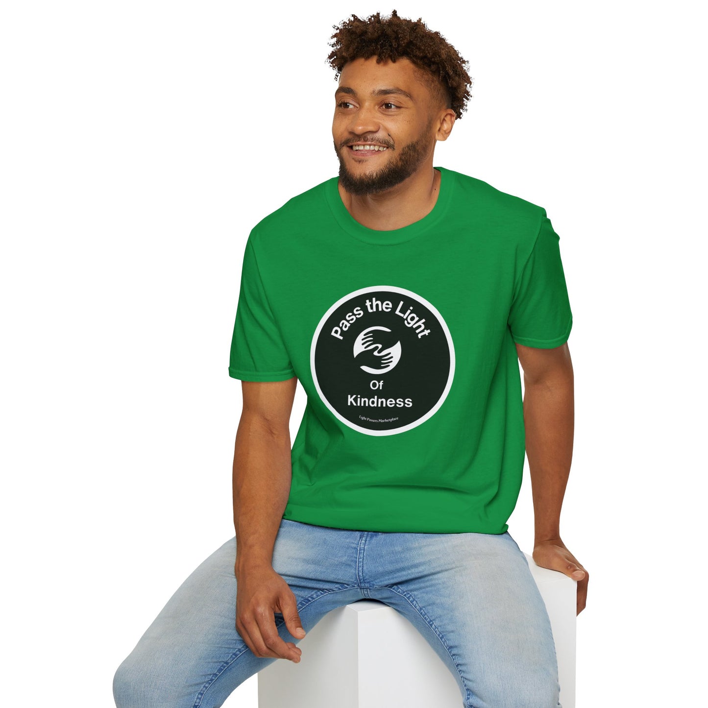 A man in a Pass the Light of Kindness 2 hands Unisex T-shirt, sitting on a cube. Green shirt with white text and black circle. No side seams, smooth surface for premium printing.