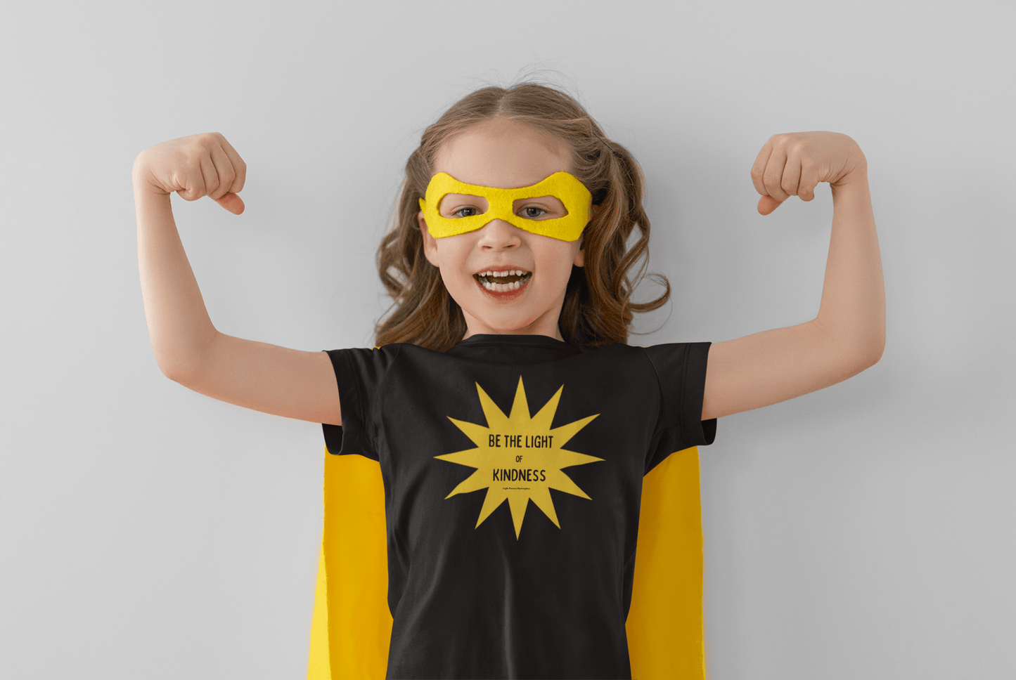 A toddler in a yellow mask and cape flexes, wearing the Yellow Star Be the Light of Kindness Toddler T-shirt. Soft, durable tee perfect for sensitive skin, with high-quality print.