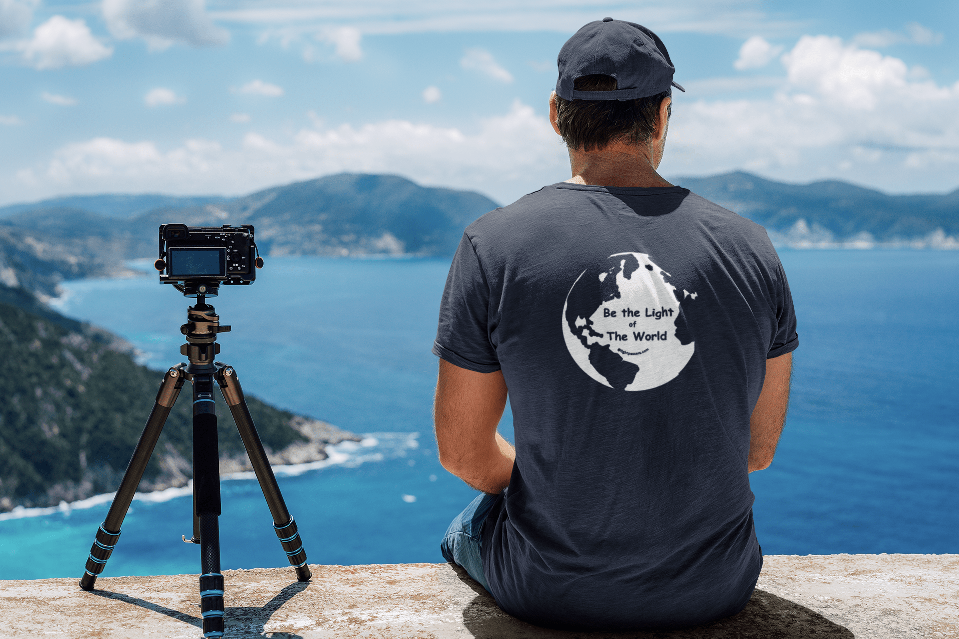 A man in a black hat sits by the ocean, wearing the Be The Light of the World Unisex T-shirt. The image showcases outdoor, clothing, and camera elements.