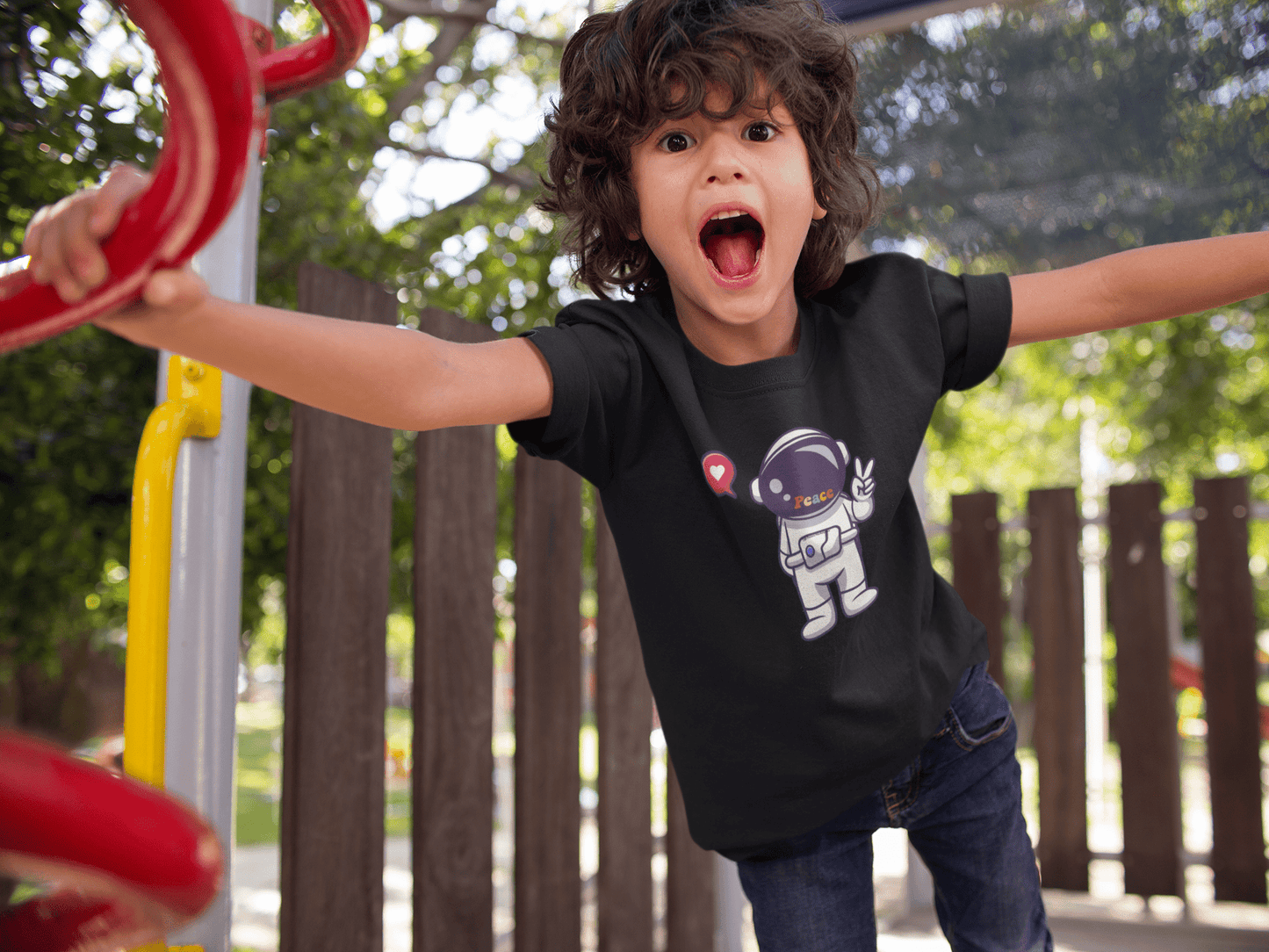 A child wearing a black shirt with a cartoon character, mouth open, in a playground. Peace Astronaut Youth T-shirt made of 100% cotton, durable twill tape shoulders, and curl-resistant collar.