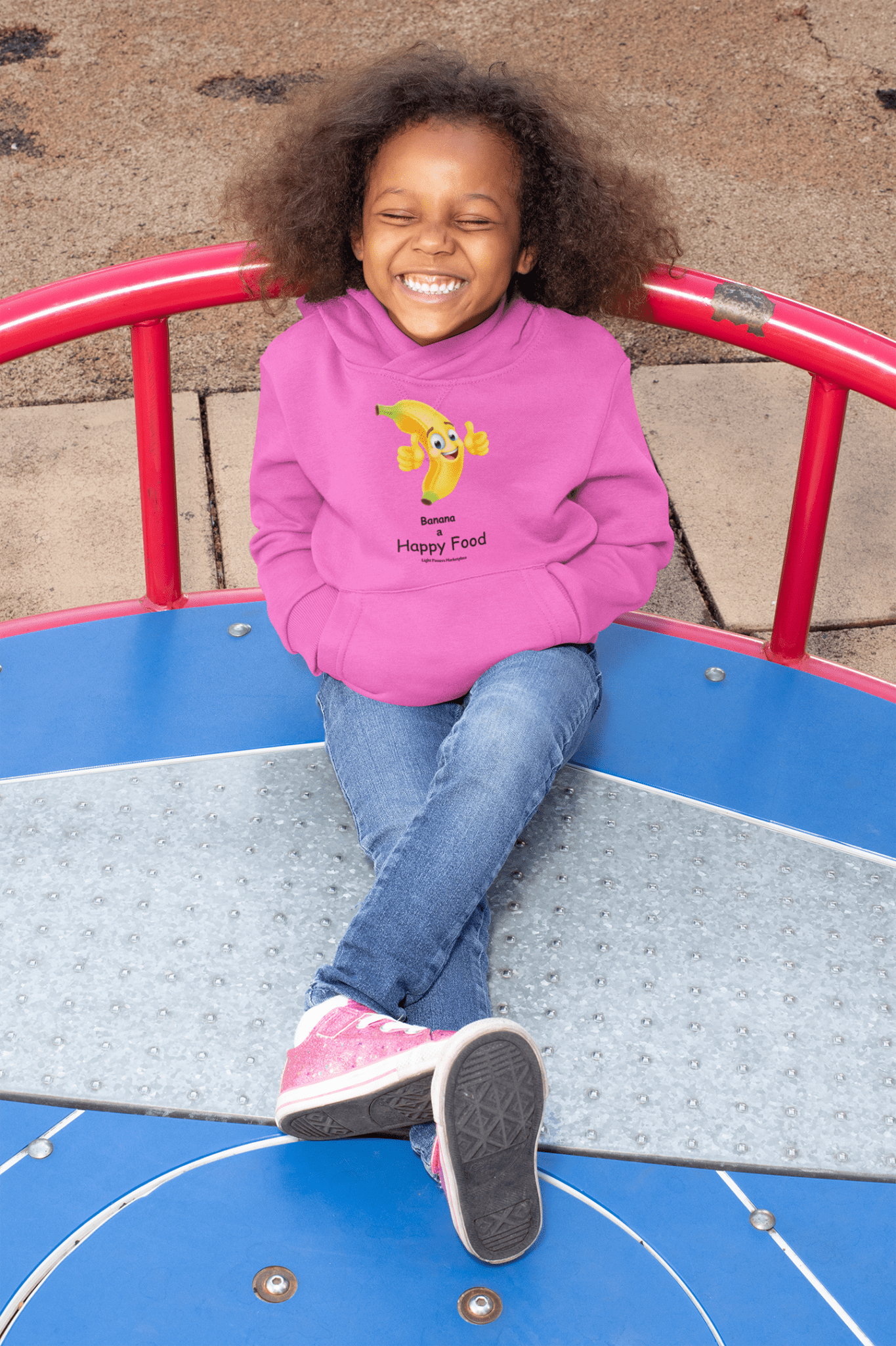 A girl sits on a merry-go-round, smiling. Product: Banana Happy Food Youth Hooded Sweatshirt. Soft fleece, kangaroo pocket, 50% cotton, 50% polyester blend.