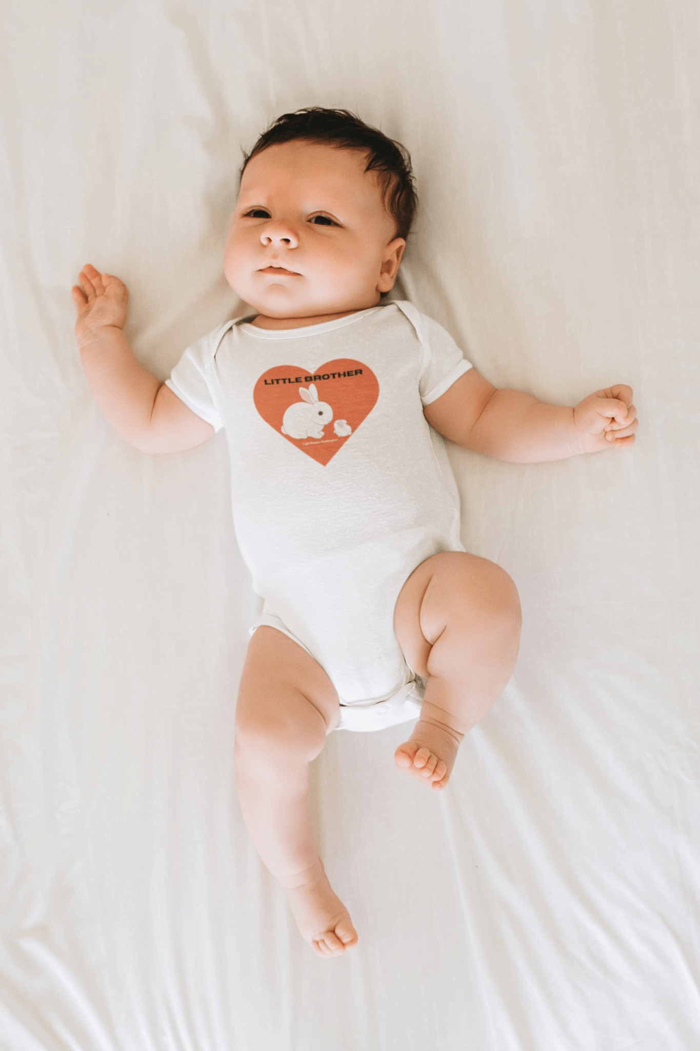 A baby boy in a Little Brother Onesie, made of 100% organic cotton, lying on a bed. Snap closure, ribbed legs for secure fit. OEKO-TEX® certified for softness and durability.