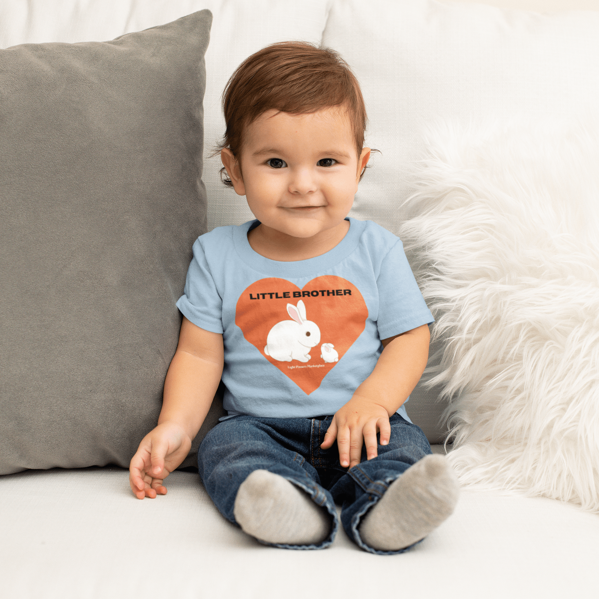A baby sitting on a couch wearing a Little Brother Baby T-shirt, showcasing comfort and durability with ribbed knitting and taped shoulders for a breezy fit.