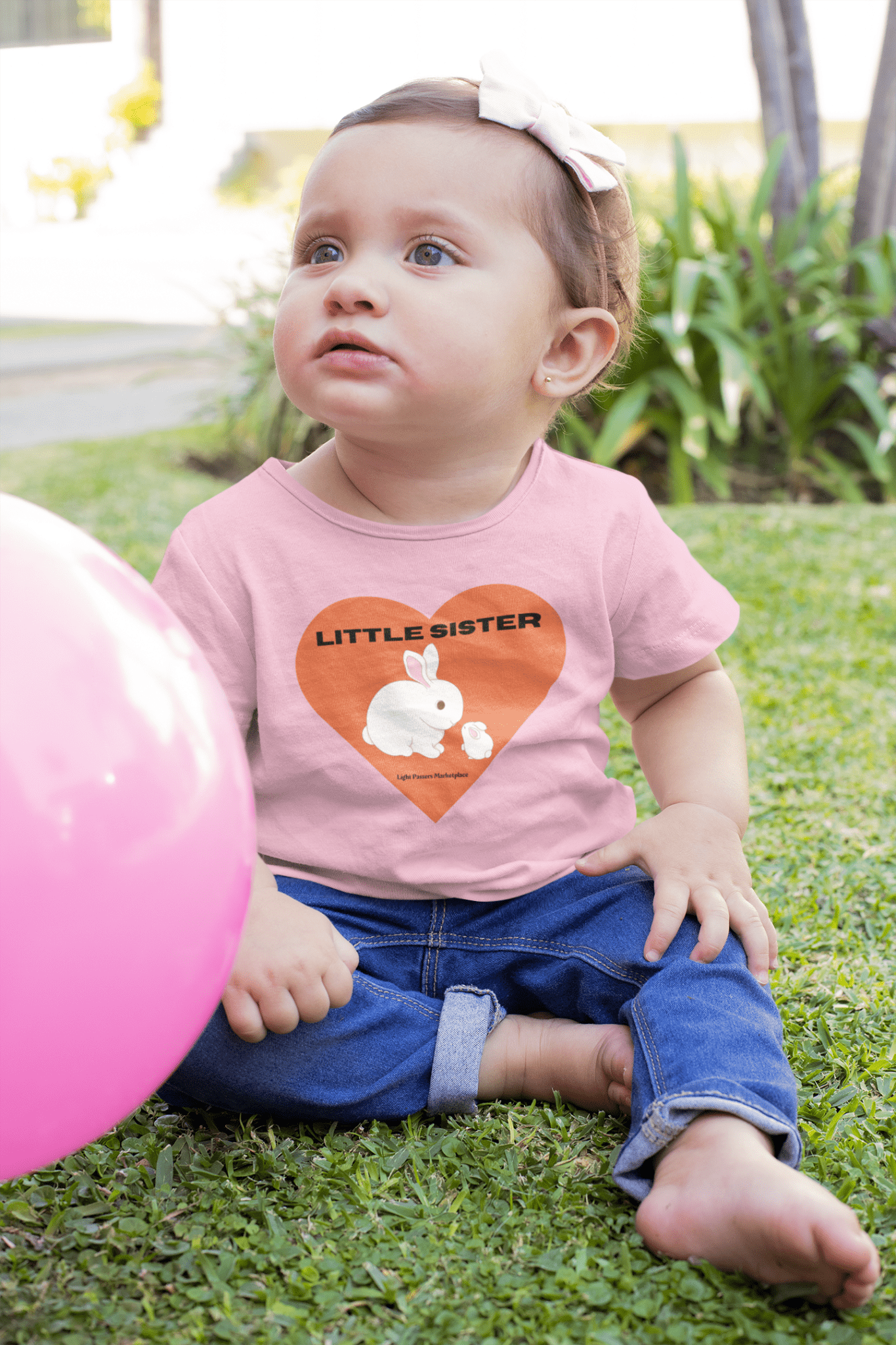 A baby sitting on grass with a balloon, wearing a Little Sister Baby T-shirt. Infant tee with side seams, ribbed knitting, and taped shoulders for comfort and durability.