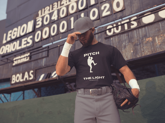 A man in a baseball uniform holding a baseball glove, showcasing the PITCH The LIGHT white lettering Unisex T-Shirt. Made of soft 100% cotton, with twill tape shoulders for durability and a ribbed collar.