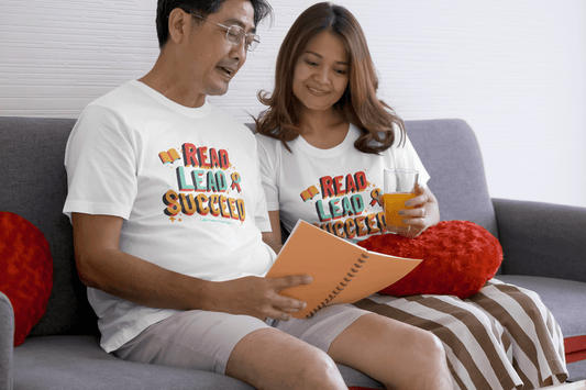 A man and woman on a couch reading, woman holding a glass of juice and a notebook. Unisex soft-style tee, 100% cotton, twill tape shoulders, ribbed collar, tear-away label.