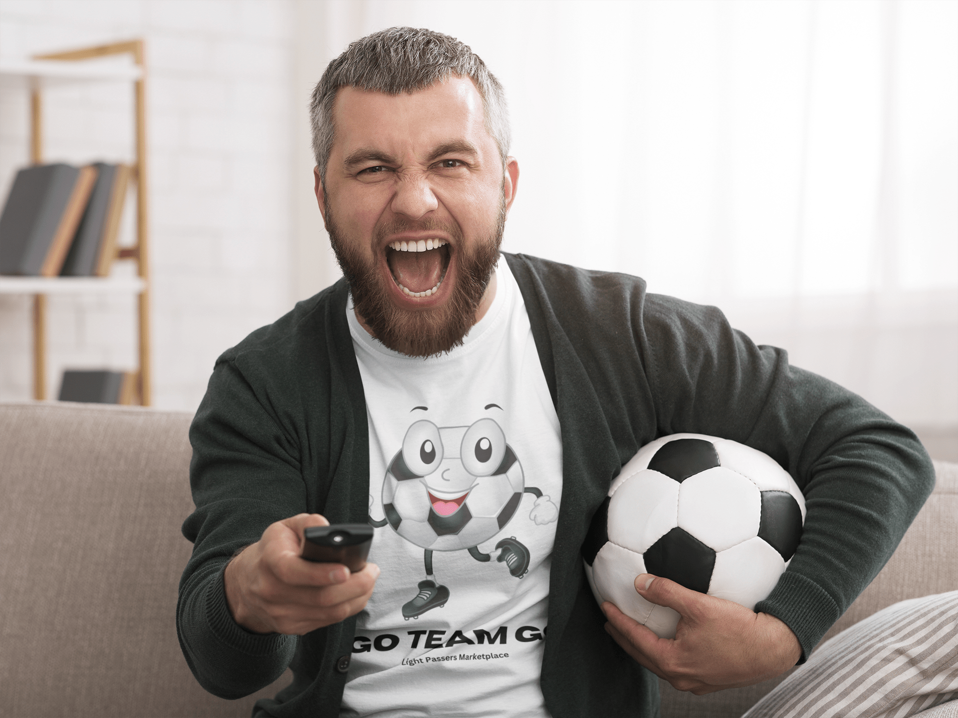 A man holding a football and remote, showcasing the Soccer Ball Guy Team Unisex T-Shirt. Made of soft 100% cotton, featuring twill tape shoulders for durability and a ribbed collar.