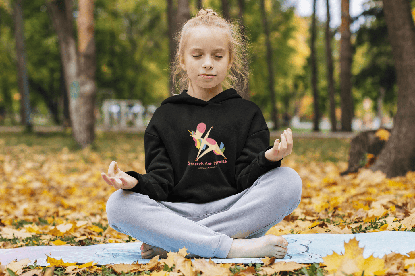 A girl in the woods sits on a rug, eyes closed, wearing a black sweatshirt with a unicorn. Product: Unicorn Stretch Youth Hooded Sweatshirt, 50% cotton, 50% polyester, kangaroo pocket, twill taping.