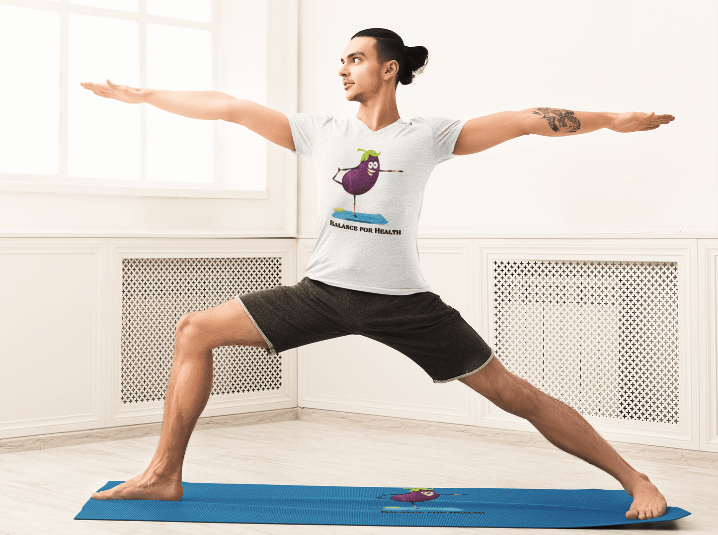 A person in a room doing yoga wearing a t-shirt with a cartoon eggplant, black pants, and a blue mat with a fish. The unisex Eggplant Balance for Health T-Shirt, 100% cotton, lightweight, with twill tape shoulders for durability.