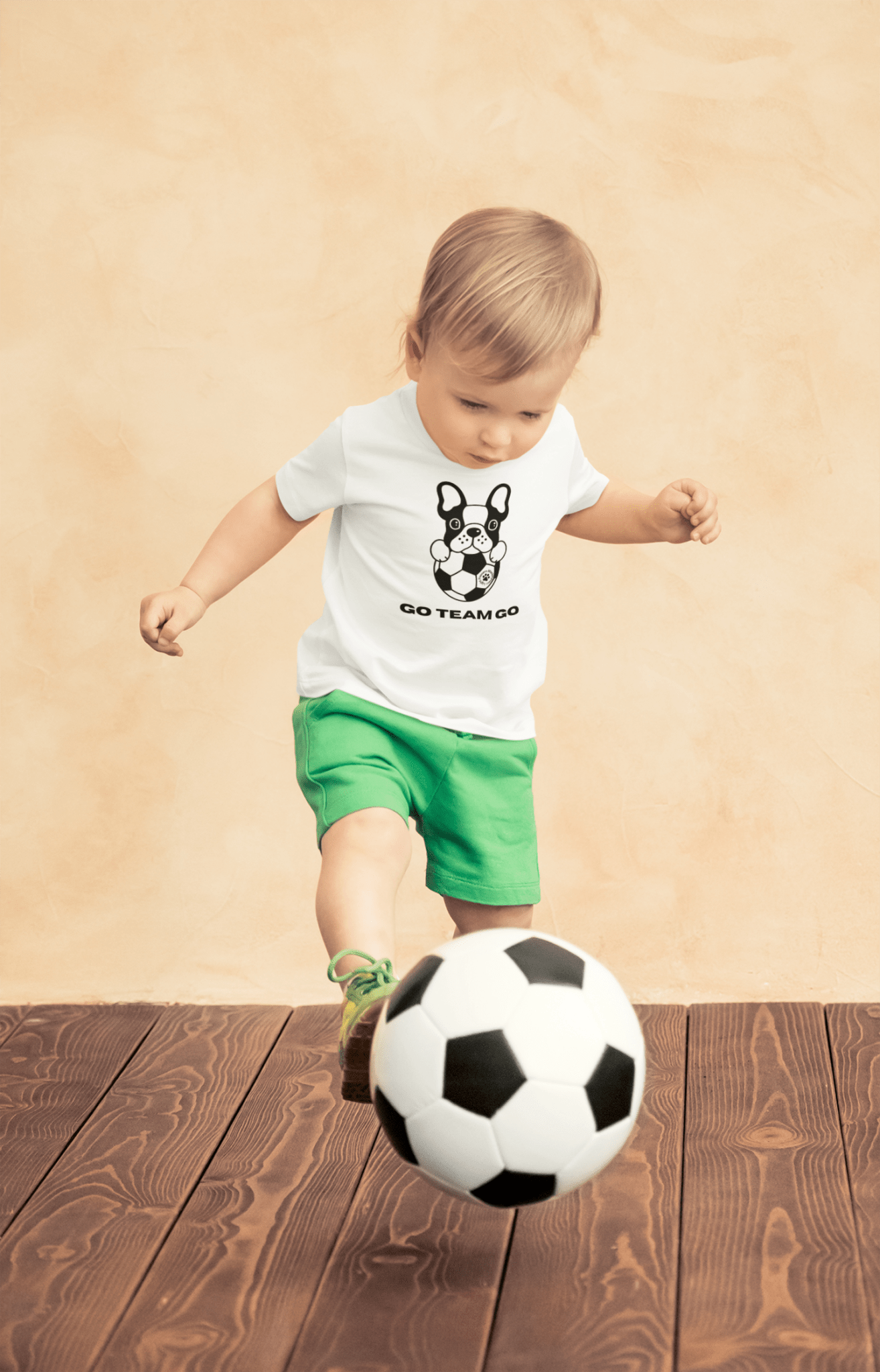 A toddler wearing a Soccer Dog Go Team Go T-shirt, kicking a soccer ball, with a close-up of the ball and a dog. Perfect for sensitive skin, 100% combed cotton, tear-away label.