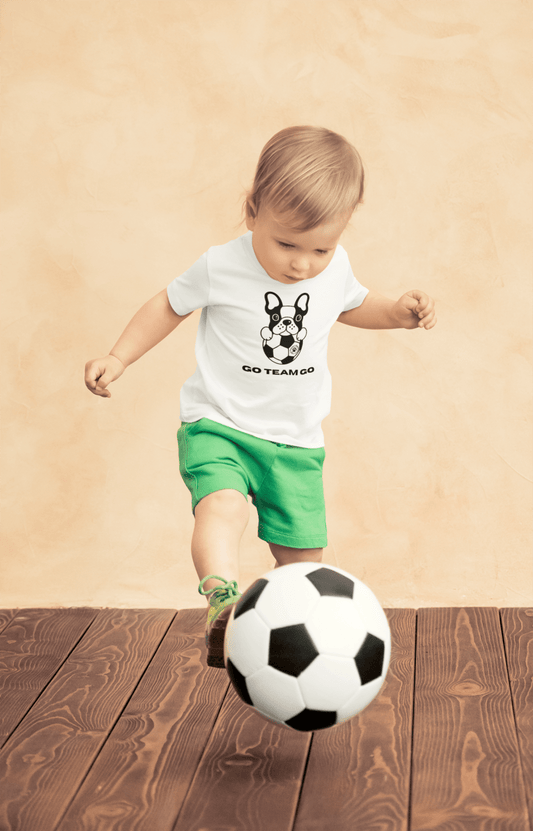 A toddler wearing a Soccer Dog Go Team Go T-shirt, kicking a soccer ball, with a close-up of the ball and a dog. Perfect for sensitive skin, 100% combed cotton, tear-away label.