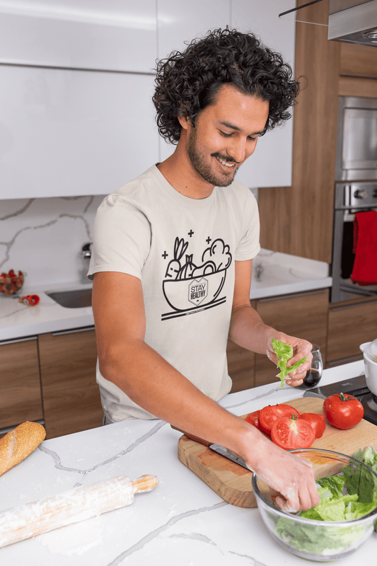 A man cutting tomatoes in a kitchen, mixing salad in a bowl, and smiling, showcasing the Stay Healthy Unisex T-Shirt's comfort and durability.