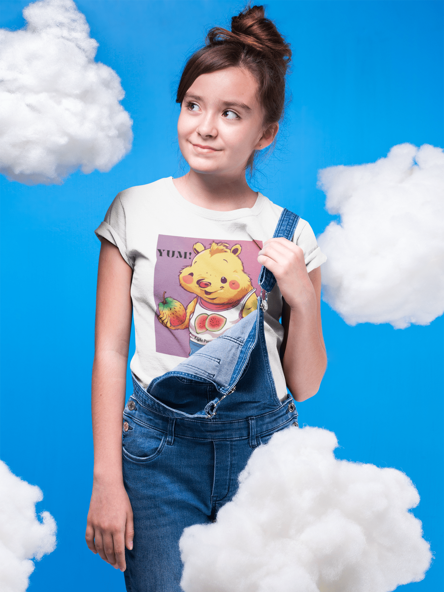 A girl in overalls holds a shirt with a cartoon bear, showcasing the Yum Strawberries Youth T-shirt. Made of 100% cotton, with twill tape shoulders for durability and a curl-resistant collar.