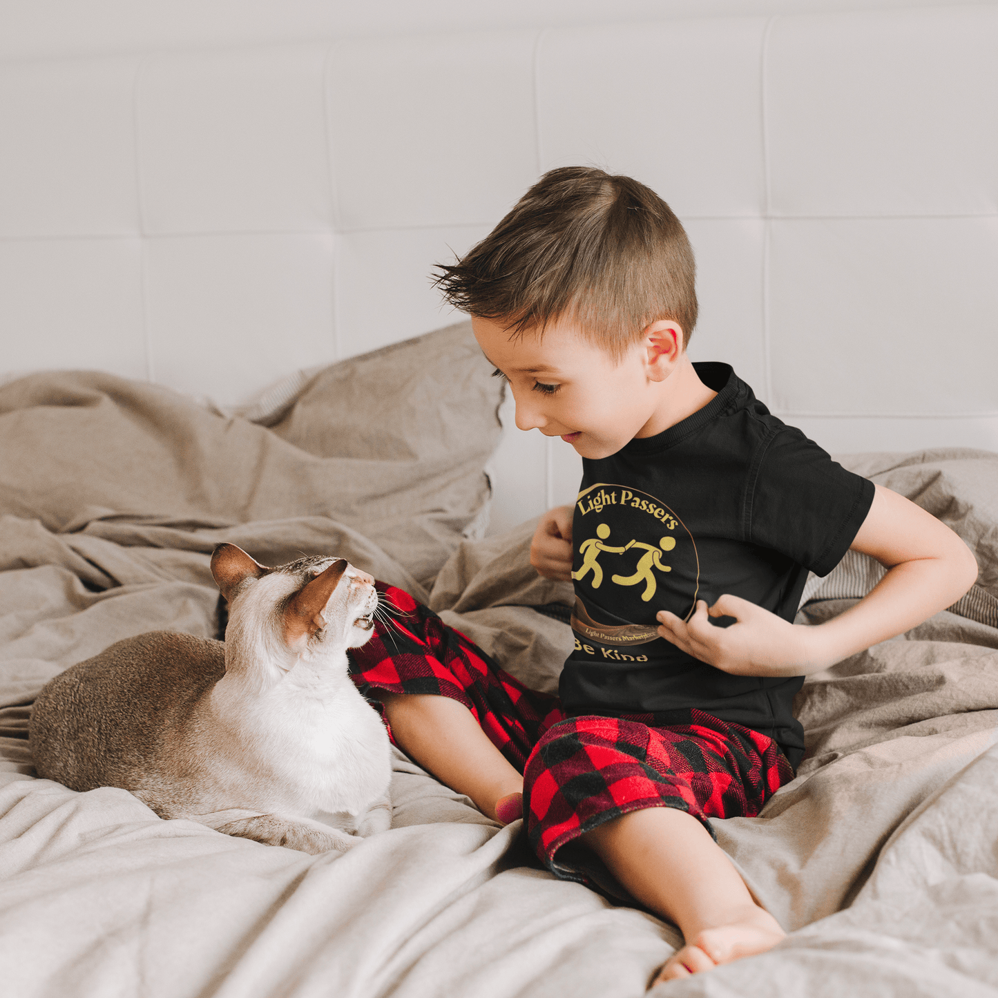 A toddler wearing a Light Passers Relay Be Kind T-shirt, playing with a cat on a bed. Soft 100% combed cotton, durable print, tear-away label, perfect for sensitive skin.