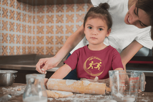 A child in a kitchen with a woman, rolling pin, mixing ingredients in a measuring cup, and hand rolling out dough. Light Passers Relay Share Toddler T-shirt, soft 100% combed cotton, durable print, tear-away label, classic fit.