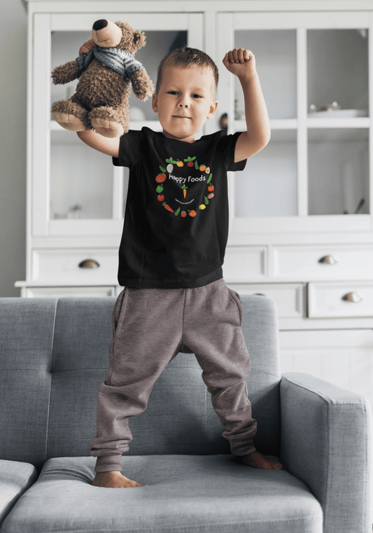 A boy in a black shirt holds a stuffed bear on a couch, showcasing the Happy Foods Ring Toddler T-shirt's softness and durability for sensitive skin.