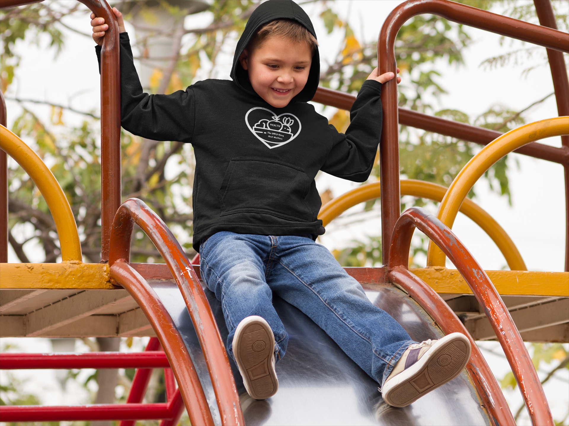 A child in a black hoodie slides down a playground slide, showcasing the Fruit Basket Youth Hooded Sweatshirt's comfort and style. Kangaroo pocket and twill taping add practicality.