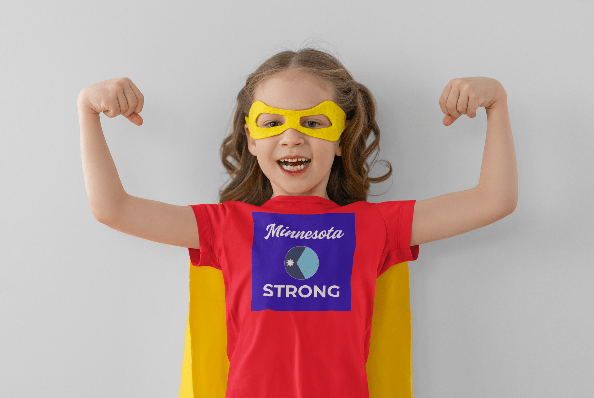 Young girl dressed as a superhero in the Minnesota Strong toddler tee, showcasing resilience and community spirit with a yellow cape and mask, smiling confidently.