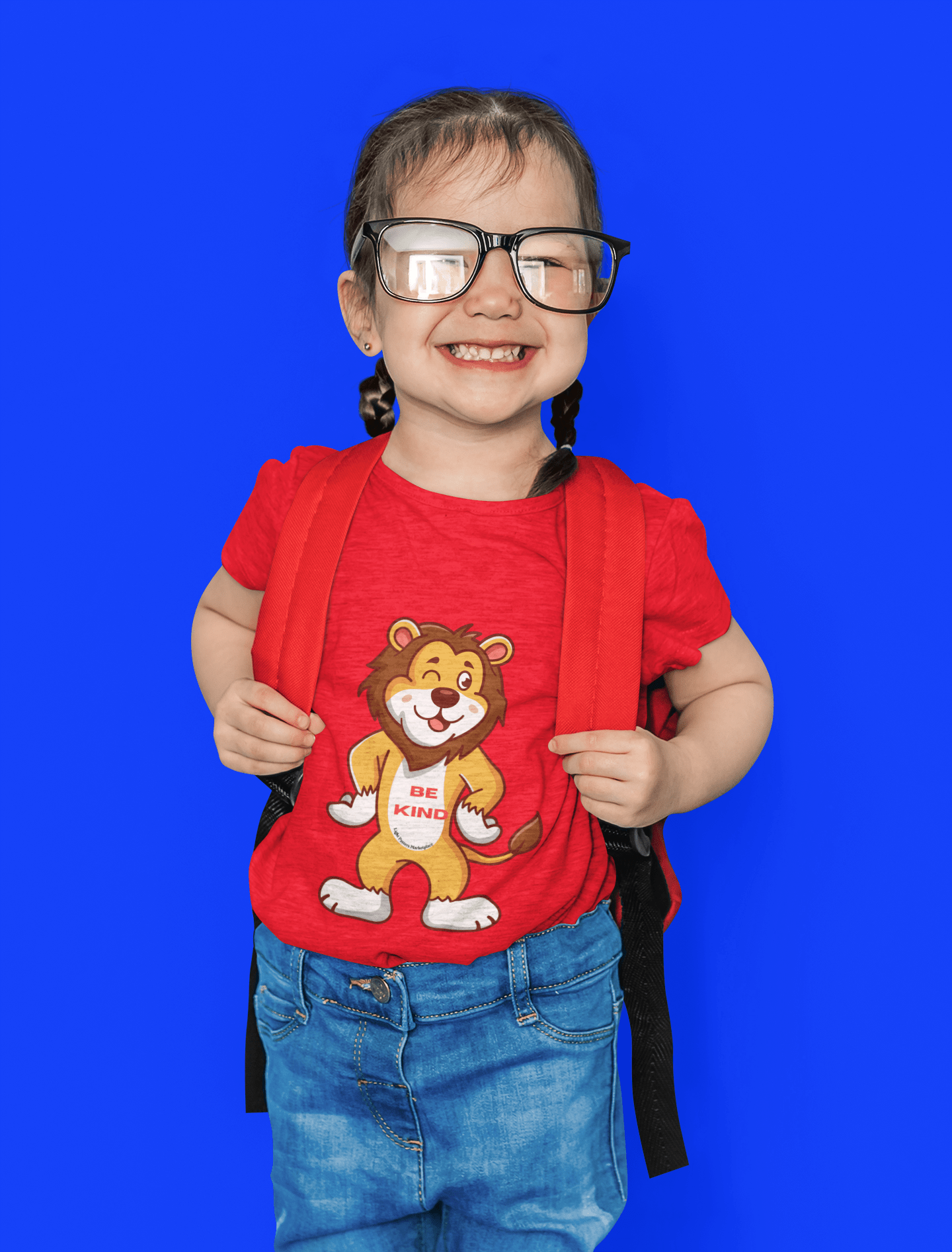 A toddler in a red shirt and glasses holds a tee with a cartoon lion print. Soft 100% combed cotton, tear-away label, perfect for sensitive skin.
