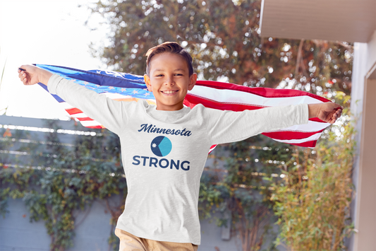Boy outdoors with arms outstretched, wearing Minnesota STRONG long-sleeve tee, holding American flag as cape. Represents youth civic courage and regional pride.
