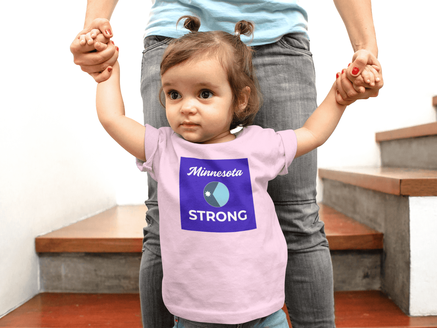 Toddler in Minnesota STRONG tee, supported by an adult, on stairs. The shirt's design reflects resilience and comfort, ideal for everyday wear.