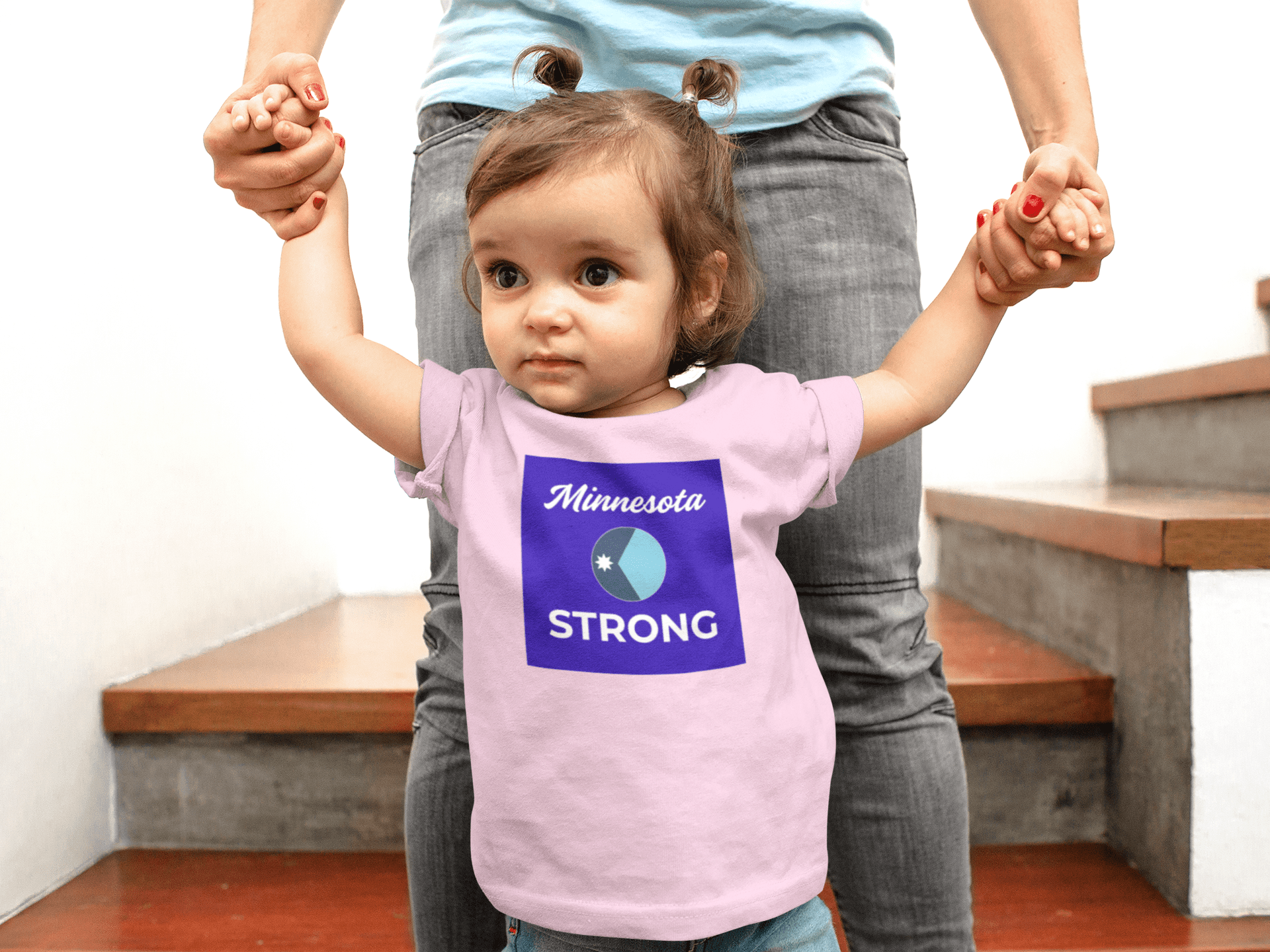 Toddler in Minnesota STRONG tee, supported by an adult, on stairs. The shirt's design reflects resilience and comfort, ideal for everyday wear.