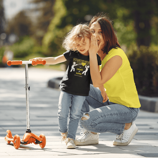 A toddler and woman on a scooter, showcasing a PASS THE LIGHT of LOVE Gold Heart Toddler T-shirt. Soft, durable tee in 100% combed cotton, light fabric, tear-away label, classic fit.