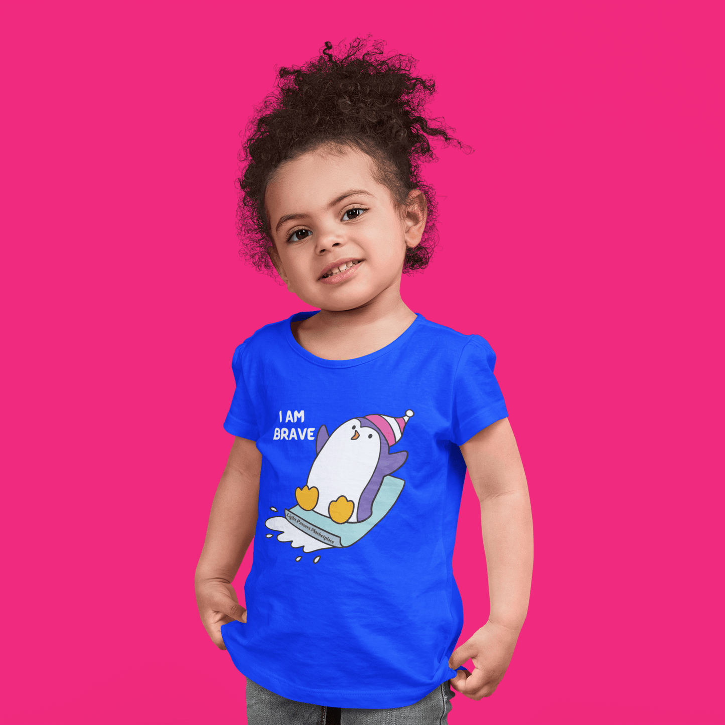 A toddler wearing a Brave Penguin tee, featuring a girl with curly hair in a blue shirt. Soft 100% combed cotton, durable print, tear-away label, classic fit.