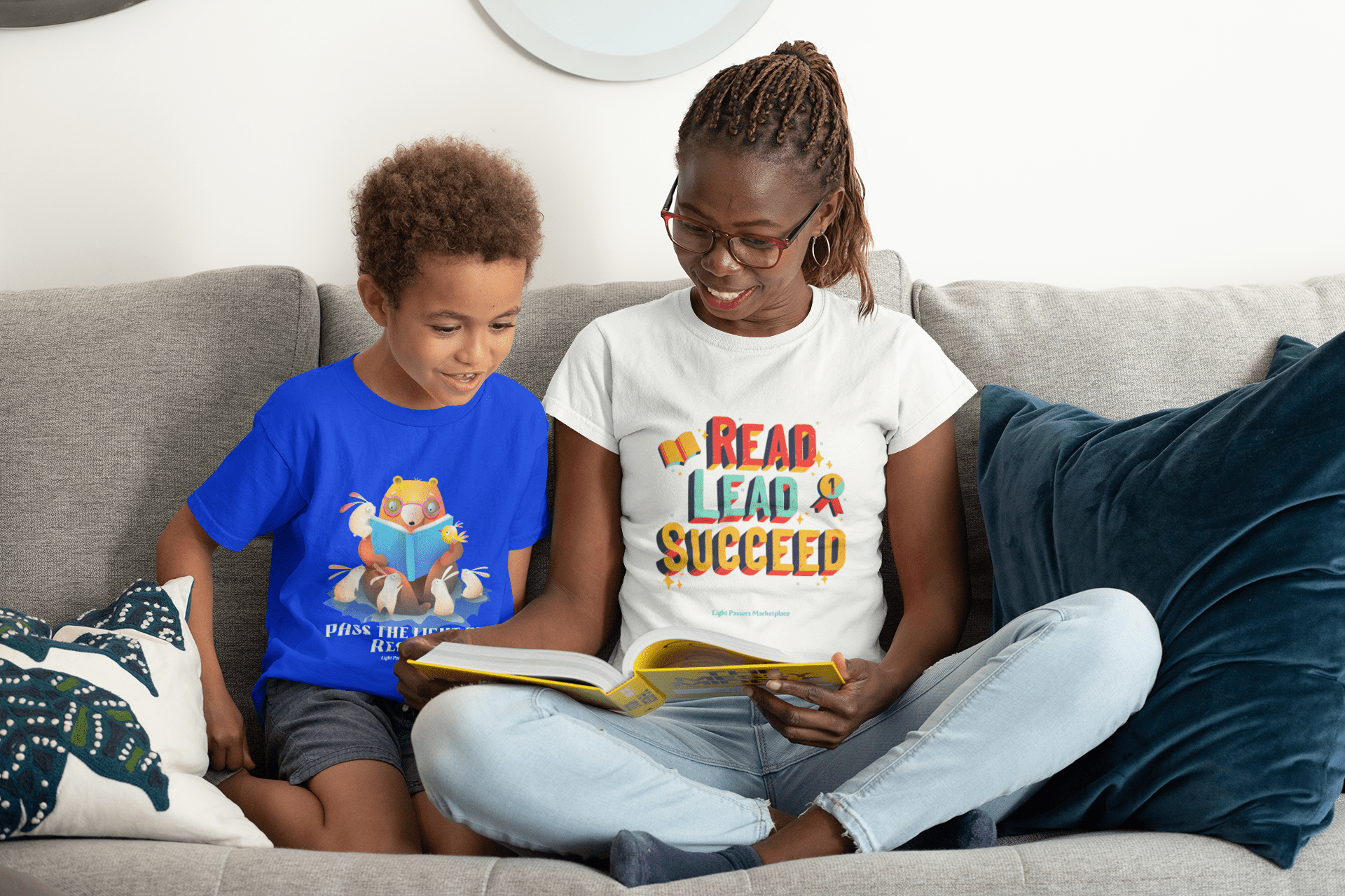 A woman and a boy on a couch reading a book, showcasing a Pass the Light of Reading Toddler T-shirt. Soft, 100% combed cotton tee for sensitive skin, with durable print.