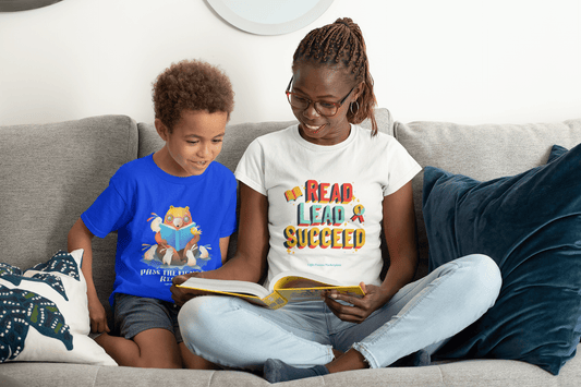 A woman and a boy on a couch reading a book, showcasing a Pass the Light of Reading Toddler T-shirt. Soft, 100% combed cotton tee for sensitive skin, with durable print.