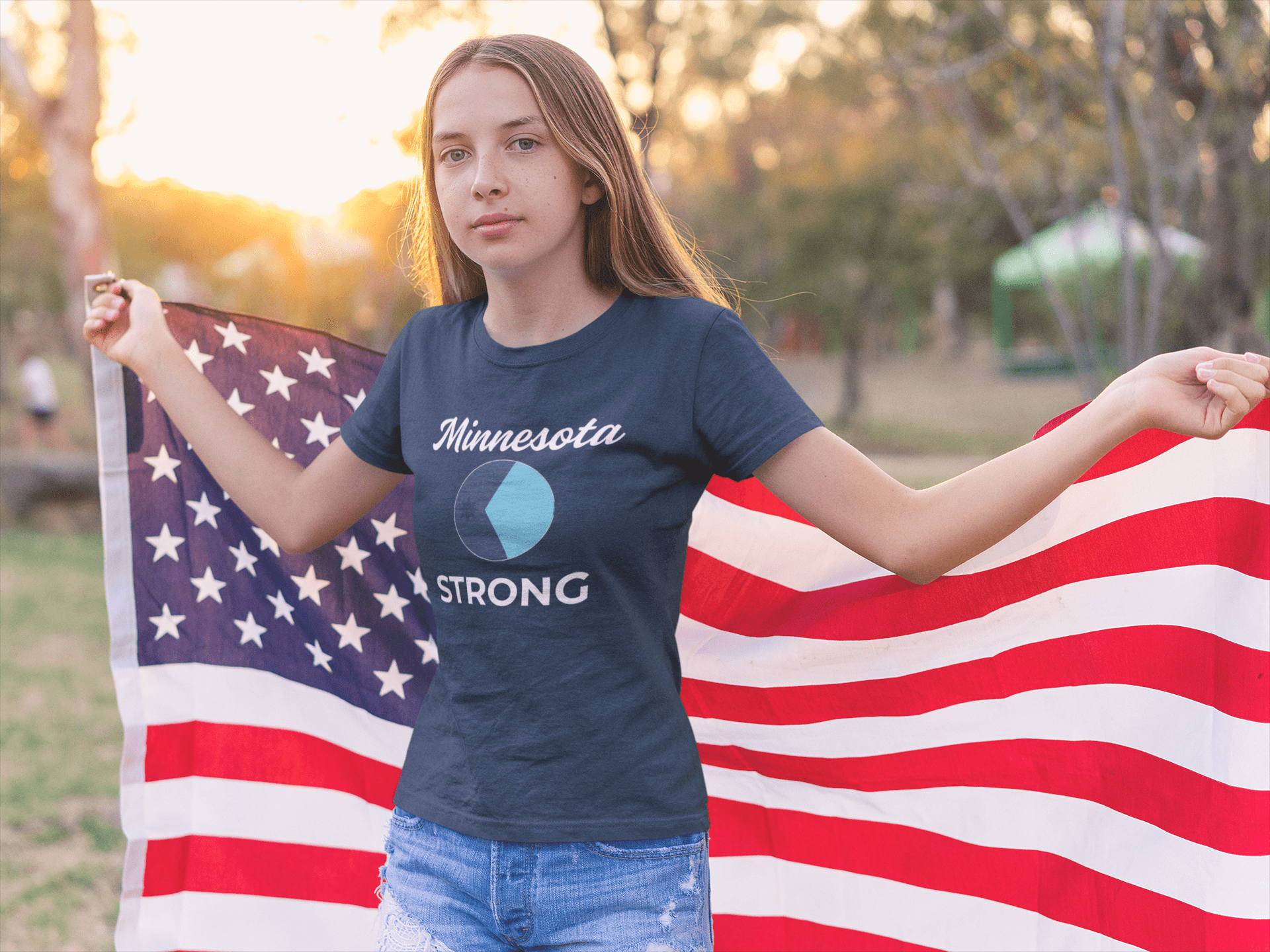 Young person holding an American flag at sunset, wearing the Love and Peace The Light Minnesota Strong Youth T-shirt, symbolizing activism and community strength.