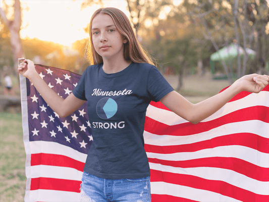 Young person holding an American flag at sunset, wearing the Love and Peace The Light Minnesota Strong Youth T-shirt, symbolizing activism and community strength.